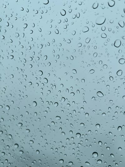 The image presents a close-up, high-angle view of numerous water droplets adhering to a transparent surface, most likely glass, such as a window or windshield. The droplets vary significantly in size, ranging from tiny specks to larger, irregularly shaped globules, and are scattered across the entire frame without any clear pattern. Each droplet refracts and reflects light, creating bright, almost iridescent highlights and distinct dark outlines or shadows, which give them a three-dimensional, spherical appearance.
Behind the water-covered surface, the background is uniformly diffused and out of focus, appearing as a soft, light grey-blue tone. This muted background suggests an overcast sky or a generally low-light, diffused environment, enhancing the clarity and prominence of the water droplets. The overall color palette is cool and subdued, dominated by blues, grays, and the bright, clear elements of the water.
The activity depicted is the presence of rain or the aftermath of rainfall, observed through a transparent barrier. The scene evokes a quiet, contemplative atmosphere, characteristic of looking out from an enclosed space during or after wet weather. No specific time of day is discernible, but the soft, even lighting indicates the absence of direct sunlight. There are no identifiable human figures, animals, or objects beyond the water droplets and the implied transparent surface and diffused background.
The OCR output provided ("C", "0", "50", "CC") does not correspond to any discernible text within the image. These characters are likely misinterpretations by the OCR engine, which may have attempted to read the shapes and outlines of the water droplets as letters or numbers. Visually, there is no text present in the image.
