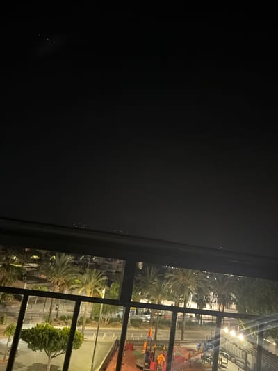 The media file is a nighttime exterior shot, oriented vertically. It appears to be taken from an indoor location, looking out through a window with dark, prominent framing bars. The scene outside depicts a street or public area with palm trees and other greenery illuminated by artificial lights. In the background, there are buildings and what looks like a playground with colorful structures and orange flags. The sky is dark, indicating it is nighttime, and there are a few distant lights visible in the very dark sky on the left side of the image. The overall mood is calm and possibly serene, with the warm glow of the lights contrasting with the darkness of the night. There is no visible text. Based on the provided location context of Sant Antoni De Portmany, Spain, the scene could be a view of a street or promenade near a beach or park area.
