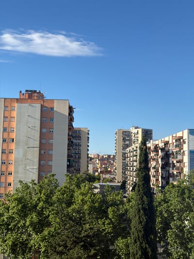 The image captures an urban view of Rubí, Spain, featuring a mix of residential architecture against a blue sky. In the foreground, a tall apartment building with a pink facade and a prominent metal wall section is visible, punctuated by rows of windows. A cluster of antennas and solar panels adorn the rooftop. To the right, lush green trees frame the composition. Further back, a succession of buildings ascend in height, showcasing various architectural styles with balconies and terraces. The clear blue sky is marked by a single, vertical cloud formation on the left side. The overall scene conveys a sunny, daytime setting in a densely populated area of Rubí.