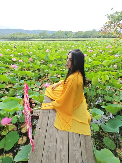 A woman in a yellow robe sits on a wooden pier surrounded by a vast field of lotus flowers and lily pads. The woman is facing away from the camera, looking towards a distant tree line and hills under a cloudy sky. She has long, dark hair and is holding a decorative, folded pink umbrella with a wooden handle. The lotus flowers are a mix of pink and white blooms, interspersed with large, green lily pads. The water is dark and reflects the surrounding foliage. The wooden pier is weathered and runs diagonally across the foreground of the image. The overall scene depicts a serene natural setting.