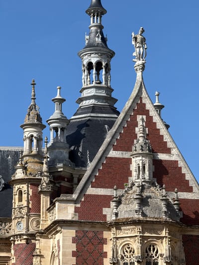 This media file captures the ornate upper facade and roofline of a historic building in Fécamp, France, set against a clear blue sky. The architecture features a striking contrast between dark red brick, arranged in intricate geometric patterns, and light-colored carved stone.
Multiple elaborate stone spires and turrets, some topped with finials, rise along the roofline, showcasing detailed columns and arches. One prominent turret is capped with a dark, possibly slate, conical roof. A large, intricately carved stone statue of a robed figure, seemingly holding an object, is perched on a decorative projection extending from the building.
The facade also includes ornate windows with delicate stone tracery and a circular clock face on one of the stone sections. The sun, likely from the upper right, casts distinct shadows, accentuating the three-dimensional detail of the numerous sculptures and architectural elements. The overall impression is one of grandeur and detailed craftsmanship, suggesting a Gothic or Neo-Gothic style. The weather is bright and sunny, indicating daytime. No discernible activity is taking place, and no visible text is present on the clock or elsewhere.
