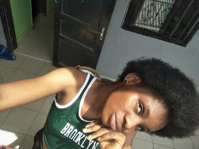 The image presents an indoor scene within the city of Cotonou, Benin, featuring a young Black woman positioned for an angled, close-up portrait. She possesses dark, afro-textured hair and is attired in a dark green sleeveless top with white trim, emblazoned with the word "BROOKLYN" in white lettering across the chest. A dark, small stud earring is present in her right earlobe. Her left hand is raised, with fingers resting against her chin. Her eyes are directed towards the camera. The background consists of a light gray painted wall on the right and upper frame. A dark brown wooden door, partially open, is visible to the left, revealing an adjacent room or hallway with bright green walls and a light-colored tiled floor. A blue object, possibly a bag, rests on the floor in this background area. Above and to the right of the woman's head, a window framed in dark wood features ornate dark metal security bars. A light-colored chair back is partially discernible behind her right shoulder. The scene is illuminated by strong direct light, originating from the left, which creates distinct highlights on the woman's skin and casts shadows within the interior space.