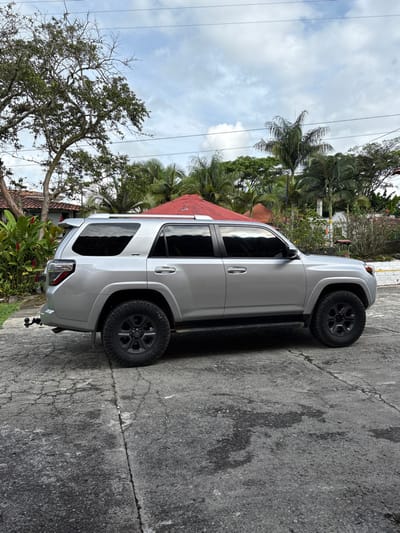 The image displays a silver Toyota 4Runner SUV, observed from its left side, parked on a cracked concrete surface in Quimbaya, Colombia. The vehicle features black multi-spoke wheels, tinted windows, and a tow hitch with a ball attached to its rear bumper. No individuals are present. The environment is characterized by lush tropical vegetation, including a large deciduous tree to the left, red-flowering plants, and numerous palm trees. In the background, structures with red-tiled roofs are visible, along with a prominent red, multi-faceted roof. Overhead power lines traverse the upper portion of the frame against a partly cloudy sky. The scene is static and appears to be illuminated by natural daylight.