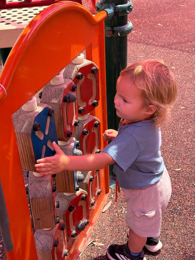 A child with blonde hair, wearing a blue shirt, beige shorts, and black and white athletic shoes, is playing on an orange and wooden playground structure. The structure features various wooden blocks with colorful tops (red and blue) and black knobs, designed for tactile exploration. The playground surface around the structure appears to be composed of small gravel. The image was taken in Kiryat Motzkin, Israel.