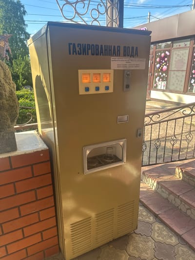This is a full shot, eye-level, outdoor photograph of a vintage soda vending machine located in Kablukovo, Russia. The machine is tan and appears to be made of metal. It has a large sign on the front with Russian text that reads "GAZIRVANNAYA VODA," meaning "sparkling water." Below the sign are three illuminated buttons with prices displayed in kopecks: "3 kopecks - plain water," "3 kopecks - water with syrup," and "5 kopecks - water with syrup." To the right of the buttons is a slot for inserting coins and a dispenser area where a glass is visible. The machine is situated on a tiled patio with a decorative metal railing and steps leading up to it. To the left of the machine is a brick wall and a large, textured rock. In the background, trees and a building with decorated windows can be seen. The lighting suggests it is a sunny day.