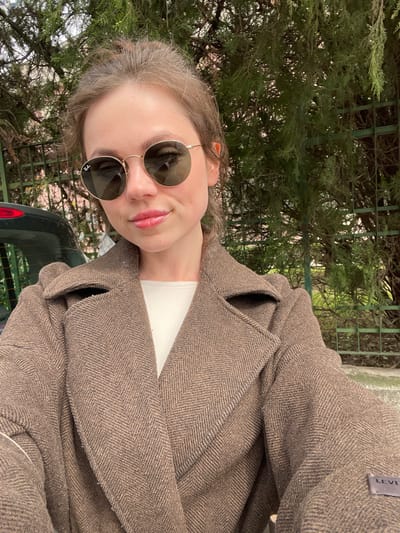 A female individual is depicted in a medium shot, facing the camera, outdoors in Cava de' Tirreni, Italy. She has brown hair pulled back and is wearing round, gold-rimmed Ray-Ban sunglasses with dark lenses, the brand logo visible on the right lens. Her lips are visible and exhibit a light pink or gloss application. She is dressed in a white inner garment beneath a thick, brown herringbone-patterned overcoat with wide lapels. A 'LEVI'S' brand label is present on the left sleeve of the overcoat. The background features dense green foliage from trees or shrubs, partially obscuring a dark green metal fence or trellis on the right. On the left, the rear section of a dark-colored vehicle, including a red taillight, is partially visible. A light-colored paved surface is visible at the bottom of the frame.