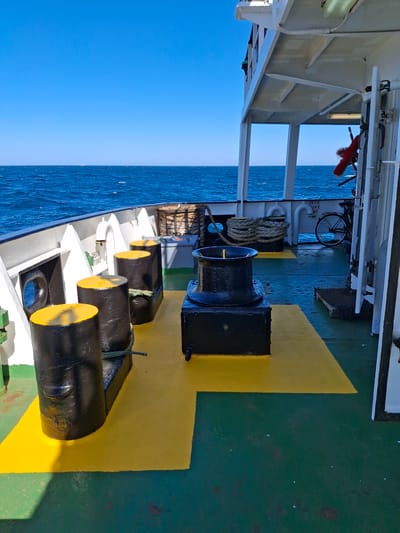 This image shows the deck of a ferry boat, likely in transit. The foreground features nautical equipment, including bollards and a winch, secured to a yellow-painted section of the green deck. In the background, the blue sky meets the blue sea. The specific location is impossible to determine from the image alone, and there is no city named Unknown in Denmark.