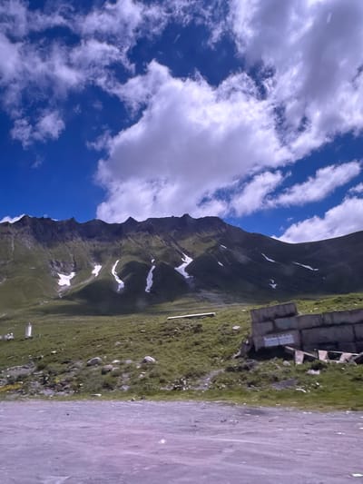 The image depicts a mountainous landscape in Gudauri, Georgia. The foreground consists of a rough, greyish-purple gravel surface. Beyond this, a grassy hillside rises towards a range of dark, rugged mountains. Patches of white snow are visible on the slopes of the mountains, contrasting with the green vegetation. The sky is a vibrant blue, dotted with numerous fluffy white clouds. On the hillside, there are several objects including a white cylindrical structure on the left and a pile of large concrete blocks on the right, with some debris scattered around. The overall scene captures a natural, rugged environment with man-made elements present.