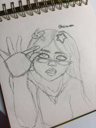 The image is a pencil sketch on paper, likely from a notebook, as indicated by the spiral binding at the top. The sketch depicts a person's face and upper body, with elements of anime-inspired style. The subject has long hair adorned with a star-shaped clip and a bunny-shaped clip. They are wearing glasses and have their hand raised, partially covering their face. The sketch is in black and white, with varying line weights to suggest form and shading. There is a signature in the upper right corner that reads "@uliixx-art". The background is blank paper. There is no information in the image to indicate a location in Velbert, Germany.