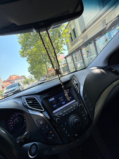 This is a point-of-view photograph taken from inside a car, likely in Brandýs nad Labem-Stará Boleslav, Czechia. The camera looks forward through the windshield and out the side windows. A rearview mirror is visible at the top of the frame, and a string of wooden beads with a cross charm hangs from it.
The car's dashboard is prominent in the foreground, featuring a center console with a media screen, various buttons, and a climate control dial. A portion of the steering wheel and the engine start/stop button are visible on the left.
Outside the car, a tree with lush green leaves dominates the left side of the scene, with its branches extending upwards. The clear blue sky is visible above the tree. In the middle ground, a silver minivan with the license plate "7AN 7764" is parked on the right side of the street. Further down the street, other cars and buildings are visible.
The buildings on the right side of the street appear to be residential or commercial, with a bus stop shelter adorned with colorful butterfly stickers and silhouettes of birds. The overall environment suggests a sunny day in a town or city.