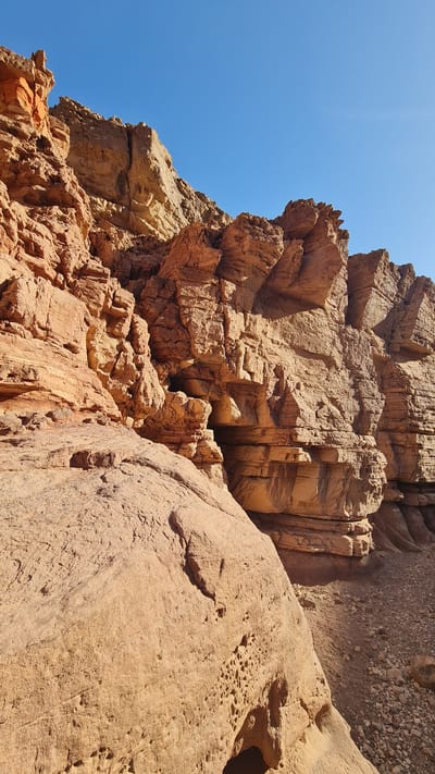 The image depicts a close-up view of a rocky terrain, likely a geological formation. The rocks are predominantly brown and orange, with varied textures, ranging from smooth surfaces to fractured and layered formations. The top of the image shows layered rocks. Some rocks have small holes. The sky is visible in the upper-left corner, appearing as a clear, bright blue. The image was taken in Hevel Eilot Regional Council, Israel.
