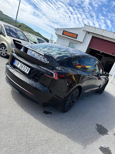 In Ørnes, Norway, a black Tesla Model 3 is parked on an asphalt lot in front of a white building with a red garage door and a sign that reads "VANOR." The Tesla has a carbon fiber spoiler and a Norwegian license plate that reads "ER 26329." A light-colored minivan is parked to the left of the Tesla. The sky is blue with scattered white clouds, and a hillside covered in green trees is visible in the background. The scene appears to be a sunny day.