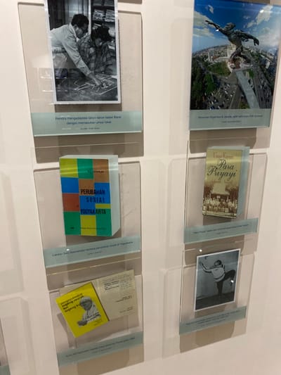 The image shows a wall display in a museum. The wall is white and the display consists of framed, black and white photographs and documents, presented in a grid pattern. The images are encased in clear acrylic frames. The images include a man in a suit directing another man, a statue of a man, a book cover, and a photograph of a man in a pose. There is text under each image in Indonesian. The location is -7.8, 110.406. The coordinates correspond to Banguntapan, Indonesia. The museum is located in Banguntapan, Indonesia. The scene is brightly lit. The museum is quiet and empty.