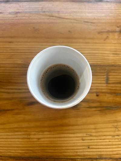 A close-up, top-down view shows a white paper cup of dark coffee, viewed from above. The coffee has a foamy layer around the rim. The cup rests on a polished wooden surface with visible grain. The image does not provide information about the city of Martinovo, Bulgaria, or any people, actions, or interactions.