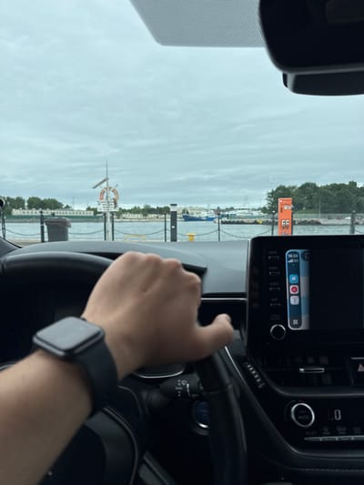 The image shows a first-person perspective from inside a car, looking out through the windshield and towards the dashboard. The driver's hand is visible, resting on the steering wheel. The car's infotainment screen is on, displaying a phone interface with icons for phone, music, maps, and messages. A digital clock on the screen shows 14:34. Outside the car, a harbor scene is visible under an overcast sky, suggesting cloudy weather. Boats are docked in the water, and a distinctive orange sign with some symbols is present on the left side of the frame. In the distance, buildings and trees line the waterfront. There is a life preserver ring visible on a pole near the water. The scene is likely taking place in Gdańsk, Poland, given the provided location context. The overall mood appears to be neutral, with no strong emotions conveyed.