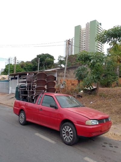 A red pickup truck is parked on a paved road in Cuiabá, Brazil, with a stack of brown wicker chairs in its cargo bed. The truck is facing towards the right side of the image. In the background, there are several buildings, including a tall, multi-story building with green accents and many windows. Trees and vegetation are visible around the buildings, and a brick wall and stacks of bricks are present in the midground. The sky is overcast and white. The scene appears to be in an urban or semi-urban environment.