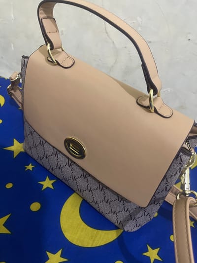 A high-angle, close-up shot captures a beige handbag with a textured, patterned fabric insert. The handbag has two top handles, both made of beige leather and attached with gold-colored hardware. A prominent gold clasp with an engraved logo is visible on the front of the bag. The patterned fabric features a repeating geometric design in grey and black. The handbag is placed on a blue fabric surface decorated with yellow crescent moons and stars. To the left of the handbag, a light-colored, textured surface, possibly a wall or floor, is visible. The scene appears to be indoors, with no indication of time of day or weather. The image is well-lit, focusing on the details of the handbag. No people are visible in the image. The location context is Anggaswangi, Indonesia.