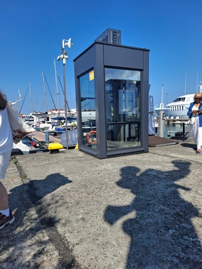 A woman walks along a concrete pier with a large, dark gray modern structure with glass walls in the foreground. She is partially visible on the left, wearing a white shirt, white shorts, and brown sneakers, carrying a patterned pillow. A second woman, further to the right, wears a blue jacket, white pants, and sunglasses, and is looking at her phone. A large white yacht is docked behind them, and numerous sailboats are moored in the background under a clear blue sky. The scene appears to be a marina in Guzeripl', Russia, given the context provided. Shadows of the two women are cast on the pier.