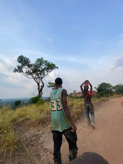 An outdoor scene in Asaga Amangwu, Nigeria, depicts two individuals walking along a dirt path on an elevated, sparsely vegetated landscape under a cloudy sky. In the foreground, facing away from the camera, a male individual wears a light blue sleeveless jersey with "HILL 33" printed in yellow and white on the back, and dark, soiled trousers. His left hand is visible and appears clenched. Further along the dirt path, a second male individual, bare-chested and wearing dark jeans, walks away from the camera. This individual carries a red plastic jerry can on their head, supported by both raised arms, and has a red cloth or bag on their back. To the left, a prominent tree with green foliage stands against the horizon, which reveals a distant, hazy panoramic view of a lower-lying area. To the right, a reddish-brown rocky outcrop is visible. The ground is dry earth with patches of tall, dry grass.