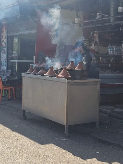 The scene depicts an active outdoor food preparation area in Boufakrane, Morocco. In the central foreground, a stainless steel commercial counter on wheels supports seven terracotta tagine pots with conical lids, arranged in a row. Significant white and blue-tinged smoke or steam emanates from the tagines and the cooking surface beneath them. An adult male, with a beard and wearing a dark cap and top, is positioned behind the counter, observing or tending to the cooking process.

To the left of the counter, the entrance to a building is visible, featuring a partially rolled-up metal security shutter. A sign inside reads "SALLE CLIMATISEE" (Air-conditioned Room). Orange plastic chairs and a table are visible near this entrance, suggesting a dining area. On the far left, a vertical menu board is partially visible.

To the right of the counter, within what appears to be another section of the establishment, large cuts of meat hang from hooks. A shelf holds multiple bottles of bottled water. Overhead, industrial-style fixtures are mounted. The ground surface in the foreground is asphalt or concrete, with a strong shadow cast by the counter under bright daylight.