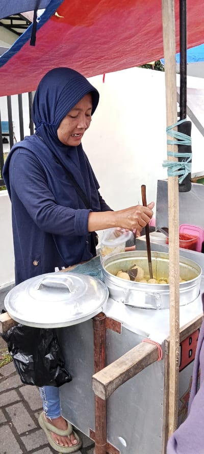 A woman wearing a blue headscarf and a dark blue jacket is standing behind a food cart in Banjaran, Indonesia. The cart has a large silver pot with a lid, a wooden armrest, and a red and white sign. The woman is using a wooden stick to stir something in the pot. There are a few other items on the cart, including a small plastic container and a black trash bag. The woman is standing on a brick walkway. The coordinates (latitude: -7.046, longitude: 107.58) correspond to the location of Banjaran, Indonesia.
