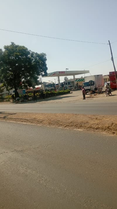 A Rubis gas station stands across a road. Several trucks and cars fill the station. A large tree is positioned on the left side of the image, next to the road. People are visible standing near the pumps and outside the station. A Coca-Cola advertisement stands near the station to the right. The sky is clear.