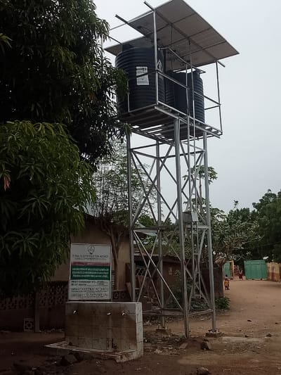 The image depicts a solar-powered water system in Biliri, Nigeria. The system consists of two black water tanks elevated on a silver metal tower, topped by solar panels. At the base of the tower is a concrete structure with multiple faucets, presumably for public water access. A sign indicates the project was commissioned by the Rural Electrification Agency for the construction of solar street lights and solar-powered boreholes in the Balanga Billiri Federal Constituency of Gombe State. The project commenced in 2019 and lasted for three weeks. The sign also lists the client as Hon. Altisa JC (PhD) and the contractor as Lorgbash Investment LTD. In the background, a dirt road leads past a house and a green gate. Several trees and foliage are visible, as well as a few people walking down the road in the background. The sky is overcast.