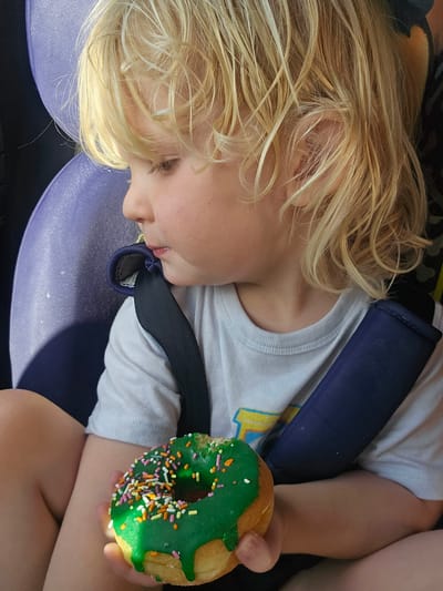 A young child with blond hair lies in a car seat, secured by a seatbelt, holding a donut with green frosting and colorful sprinkles in their left hand. The image was taken during the day. The location is unknown, thus the image cannot be confirmed to be in Key Largo, United States of America.