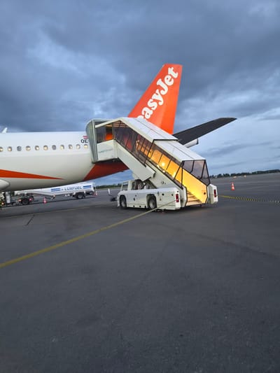 The image captures an EasyJet airplane parked at the Luxembourg Airport in Niederanven, Luxembourg. A mobile stairway is connected to the plane, illuminated from within. A fuel truck, bearing the word "LUXFUEL", is also positioned nearby. The backdrop reveals an overcast sky and a paved airport apron with painted yellow lines and orange cones.