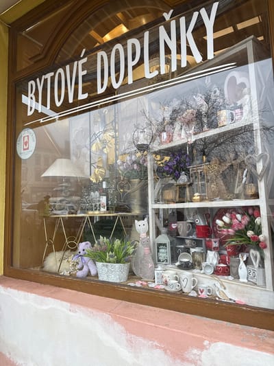 The image depicts a shop window display located in Znojmo, Czechia. The large glass window is framed in a brown-gold material, featuring an arched top. White vinyl lettering across the upper part of the window spells "BYTOVÉ DOPLŇKY" (HOME ACCESSORIES). A circular "Official Stockist Yankee Candle" sticker with a heart icon is visible on the upper left glass.

Inside the shop, a variety of home decor items are arranged. On the left, a slender gold-colored console table with an X-shaped base holds a large white lampshade, a wine bottle, two wine glasses, and other small decorative items. Behind this table, gold ginkgo leaf wall art is visible. On the floor beneath the table, a small purple teddy bear sits beside a white fluffy rug or pillow.

To the right, a white multi-shelf display unit is filled with numerous items. These include various ceramic mugs, some with heart designs, artificial floral arrangements featuring tulips, lavender, and purple flowers, decorative lanterns, small statues of cats, angels, and birds, and assorted candles and giftware. The overall aesthetic suggests a focus on romantic or festive themes, particularly with the prevalence of hearts and red accents.

Reflections on the window surface reveal elements of the street scene outside. Bare tree branches, parts of buildings, and what appears to be a distant person or vehicle on a street are faintly discernible, indicating an urban setting. The wall directly below the window is painted in two horizontal sections: a rough white texture on the upper portion transitions into a light peach or pink color below.