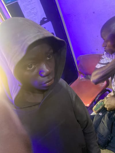A medium close-up shot shows a young Black male wearing a dark hooded sweatshirt, with the hood pulled up, obscuring the top of his head. His face is turned slightly to the right and his eyes are looking forward. Purple light illuminates the scene from the right. In the background, a red plastic chair is visible, and to the right of that, another Black male, partially visible, is wearing a white shirt with a repeating pattern of interlocking "GG" logos. A notice with text is mounted on the wall behind the young male. The scene appears to be indoors, possibly in Barakin, Nigeria.