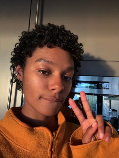 A person is centered in the frame in front of a refrigerator making a peace sign with their right hand. They have dark curly hair and a light complexion. They are wearing an orange fleece zip-up sweater. The background includes a stainless steel refrigerator and appliances. The location is unknown and cannot be determined to be in Champs-sur-Marne, France.