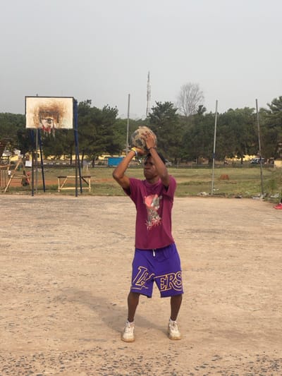 A young male is depicted mid-action on an outdoor, unpaved court in Uturu, Nigeria, preparing to shoot a basketball. He stands center-frame, facing slightly left, holding a dark, worn basketball with both hands elevated above his head. He wears a dark maroon t-shirt with a graphic print, purple athletic shorts featuring "LAKERS" in yellow lettering, and white athletic shoes. A multicolored wristband is visible on his left wrist. His shadow extends to the lower left on the light brown, uneven dirt or sand court. In the background to the left, a severely rusted basketball hoop with a white backboard and red rim is visible. The horizon is lined with green trees. A tall, slender metallic communication tower is prominent above the trees on the right side of the frame, accompanied by several vertical metal poles. The sky is a uniform, hazy light gray.