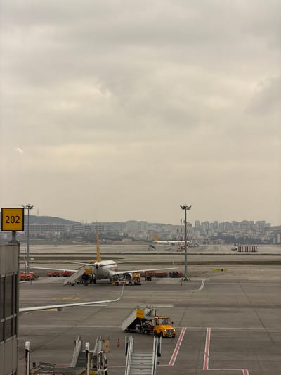 An overhead view captured from inside an airport terminal depicts the tarmac at Sabiha Gökçen International Airport, located in the Pendik district of Istanbul, adjacent to Tuzla, Turkey. The sky is uniformly overcast.

In the mid-ground, a white commercial passenger jet, identifiable as a Pegasus Airlines aircraft by its yellow tail and fuselage branding, is parked on the concrete tarmac. An articulated passenger stair unit is deployed at the aircraft's rear port-side door, while numerous red ground service carts, presumably for baggage or cargo, are clustered around both wings and the rear fuselage. A yellow ground power unit is visible near the aircraft's underside. A yellow mobile passenger stair vehicle is positioned to the right of the central aircraft.

In the immediate foreground, partially obscured, are the upper sections of two stationary passenger stair units. To the left, a prominent yellow rectangular sign with black numbers "202" is mounted on a grey pole. Multiple tall light poles with arrays of luminaires are scattered across the tarmac.

Further in the background, another white commercial passenger jet with a yellow tail is visible on a parallel taxiway. The distant background reveals a dense urban skyline composed of numerous multi-story buildings, sprawling across sloping terrain and culminating in hills under the overcast sky. A faint, small aircraft with two contrails is discernible in the upper left portion of the cloudy sky. No identifiable individuals are visible in the scene.