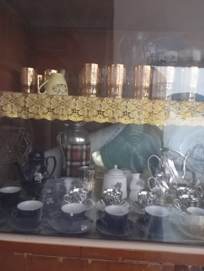 A glass cabinet displays an assortment of tea sets and glassware. On the top shelf are decorated gold-rimmed glasses and a small pitcher. Below, various tea sets are arranged, including blue cups and saucers, silver-toned sets, a plaid-patterned thermos, and sugar bowls. The cabinet appears to be of vintage design, with wooden framing and a slightly distorted view through the glass.