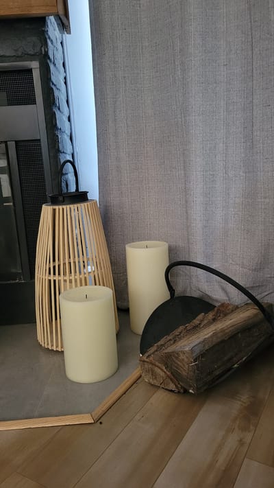 This is a photograph of a rustic indoor setting, possibly a home or a decorative display. The scene is dominated by natural materials and a focus on warm, ambient lighting.
In the foreground, a woven wooden lantern is positioned on a dark surface, likely a hearth or a low table. Inside the lantern, and also beside it, are two large, cream-colored pillar candles. These candles appear to be artificial, given the sharp wick visible in one. To the left of the lantern, a grey textured fabric hangs, creating a backdrop and suggesting the presence of a blind or a curtain.
Above and slightly to the right of the candles, a piece of raw, split firewood rests on a dark, circular base, perhaps a candle holder or part of a decorative arrangement. The wood is rough and shows the natural grain and texture of timber. The floor in the background is made of light-colored wooden planks laid in a herringbone pattern, indicating a deliberate interior design choice.
To the left and further back, a section of what appears to be a wall constructed of dark bricks is visible, with a metal grate or ventilation cover embedded in it. Above this brickwork, a bright white light suggests a window or an opening to the outside.
The overall impression is one of cozy, perhaps autumnal or wintery, decor. The lighting is soft, with the candles being the primary light source in the immediate vicinity. The time of day is not definitively ascertainable, but the indoor setting and the presence of candles suggest it might be evening or a dimly lit interior. There is no visible text in the image. The location context of Terrebonne, Canada, doesn't provide specific visual cues within the image itself.