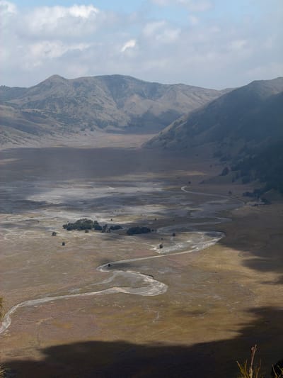 This is a high-angle shot of a vast, arid plain in Cemorolawang, Indonesia, likely part of the Tengger Caldera. The landscape is characterized by rolling, barren hills in the background, with softer, eroded slopes in the midground. A dry, winding riverbed snakes across the plain, marked by lighter-colored sediment, suggesting periodic water flow. Sparse vegetation, appearing as patches of low-lying grasses and a cluster of trees in the mid-left, dots the otherwise desolate terrain. The ground is a mix of sand, dust, and dry soil, with visible tracks or pathways suggesting human or animal passage. Shadows cast by unseen objects, possibly mountains or clouds, create varying degrees of light and darkness across the scene, contributing to a stark, dramatic atmosphere. The sky above is a pale blue with scattered, wispy clouds. No people or animals are visible in the frame. The overall impression is one of a remote, untamed, and somewhat melancholic natural environment.
