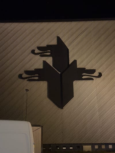 This image depicts a black, non-textual logo or symbol affixed to a gray, horizontally ribbed wall. The symbol resembles a cross-like shape with stylized, elongated appendages resembling tails, possibly reminiscent of a stylized animal or abstract design. A portion of a white vehicle is visible in the lower right corner of the image. The presence of the vehicle, combined with the context of Volketswil, Switzerland, suggests the image depicts a sign on a building in that city. The light conditions indicate it is either nighttime or a dimly lit area. A utility panel is visible in the upper right corner of the image, near the white vehicle.