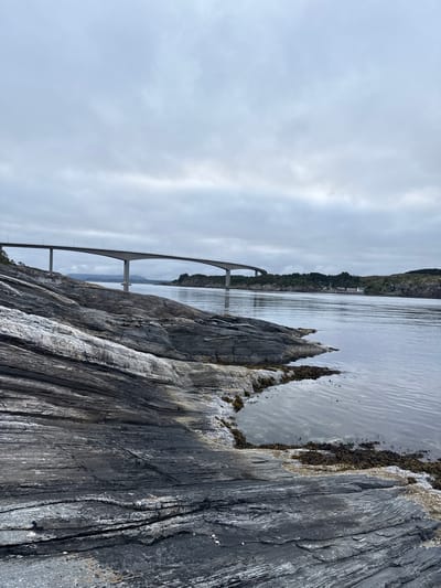 This is an image of a wide, calm body of water under a cloudy sky. In the background, a long, modern bridge spans the water, supported by concrete pillars. The bridge extends towards a hilly shoreline with scattered trees and some buildings. The foreground is dominated by dark, layered rock formations leading down to the water's edge. The water reflects the overcast sky, creating a muted and serene atmosphere. The scene is located in Rong, Norway.