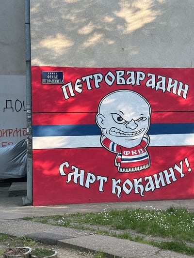 A colorful mural painted on an outdoor wall is the central focus of the image, set in what appears to be a sunny daytime environment. The mural features a bold red background with a prominent vertical stripe design of blue and white running down its center. On the left side of the mural, the word "ПЕТРОВАРАДИН" (Petrovaradin) is written vertically in large white Cyrillic letters with a black outline. On the right, the phrase "СМРТ КОКАНУ!" (Death to Kokan!) is similarly displayed vertically.

At the heart of the mural is a cartoon character: a bald, light-skinned man with a wide, toothy grin and mischievous eyes. He is depicted wearing a striped scarf in red, white, and blue, which bears the Cyrillic acronym "ФКВ" (FKV), likely representing a local football club.

To the left of the mural, a blue street sign with white Cyrillic text reads "УЛИЦА ФРАЊЕ ШТЕФАНОВИЋА" (Franje Štefanovića Street). The setting appears to be outdoors, with grey pavement visible at the bottom and to the right of the mural, alongside a patch of green grass with small white flowers. Shadows from overhead foliage are cast across parts of the wall, indicating bright sunlight. Below the mural, partially visible and inverted, are fragments of Cyrillic text, including "ДО[Л]" and "ПИР[АТЕ]". The mural conveys a strong sense of local identity, likely related to sports fan culture in Petrovaradin, Serbia.