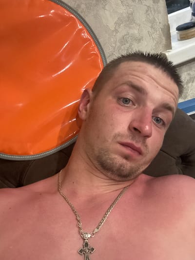 The image shows a close-up, over-the-shoulder selfie of a Caucasian male with short, spiky dark hair and blue eyes. He has a light beard and is wearing a silver chain with a cross pendant. His skin appears flushed, possibly from sunburn. He is lying down, with his upper body partially visible. To his left, a portion of a large, shiny orange object with a grey trim is visible. The background is a textured wall and a white ledge on which a small jar with a label is placed. The image appears to be taken indoors.