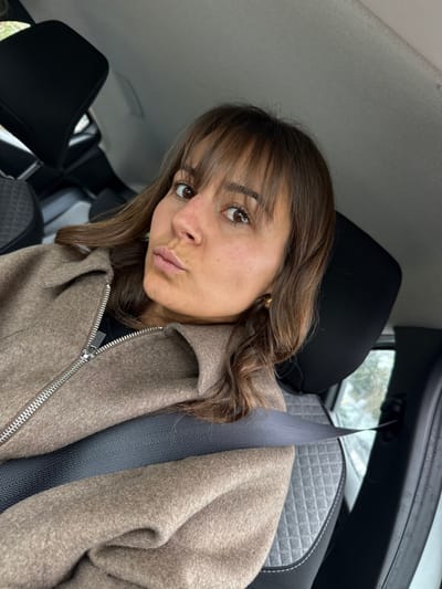 The image displays a single individual, a female with medium brown hair styled with bangs, seated in the front passenger seat of a vehicle located in Mougins, France. She is depicted from the chest up, looking directly towards the camera with a pursed-lip expression. She wears a light brown, collared jacket with a visible silver zipper, partially unzipped over a black garment. A gold-toned earring is visible in her right ear. A dark grey seatbelt crosses diagonally over her chest. The vehicle's interior features a dark grey seat with a lighter grey, diamond-quilted pattern on the seatback and cushion. A black headrest is behind her. The vehicle ceiling is light grey, and additional vehicle seats are visible in the background. Through the passenger window, a blurred outdoor environment with hints of greenery and a wet surface is discernible, suggesting ambient daylight conditions.