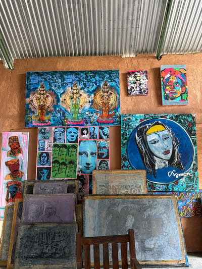This image depicts an outdoor art display in Tejakula, Indonesia, likely a stall or small gallery. The wall behind the art is made of a rough, orange-brown plaster, topped with a corrugated metal roof. A variety of artworks are arranged, with some leaning against the wall and others stacked.
Dominating the upper portion of the display is a large, vibrant blue canvas featuring two figures with multiple arms, rendered in a stylized, spiritual motif. To its right, a smaller, colorful abstract portrait of a face is hung. Below these, a collection of smaller framed pieces and canvases are arranged. One features a grid of black and white portraits, and another, a blue circular canvas with a portrait of a woman with light blue skin, is particularly prominent. To the left, a tall, narrow canvas displays two portraits in a folk art style, one with a red headdress and the other with a turban.
The lower section of the image reveals several large, rectangular screens or canvases, some appearing to be used for printing or displaying stencils. These are mostly textured with muted colors like purple, gray, and blue, with faint images or patterns visible. One large screen in the foreground features a detailed, metallic-gray image of what appears to be a dragon. A wooden chair is partially visible to the right of these screens. In the background, a sign reads "FOR SALE WORKS." The overall impression is of an artist's workspace or a market stall showcasing a diverse range of artistic expressions.