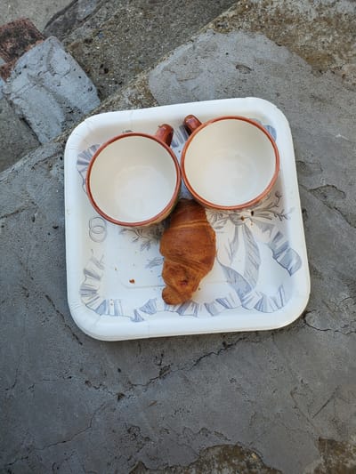 A white tray with a floral pattern rests on a cracked concrete surface, possibly a set of outdoor stairs. On the tray are two empty ceramic cups with brown rims and handles, positioned like eyes, and a croissant acting as a mouth, creating a face-like arrangement. The background shows additional concrete and brick structures, characteristic of an urban environment. The image appears to be a still life composition, with no discernible human presence or activity beyond the arrangement of the objects. The lighting suggests it was taken during the daytime.