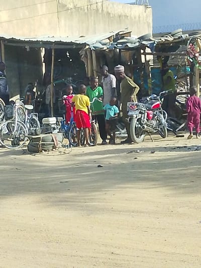 The image captures an outdoor scene, likely a bustling street or market area, under bright, possibly midday sunlight which casts distinct shadows on the dusty ground. In the background, a rudimentary stall or workshop is visible, constructed with a corrugated metal roof and supported by wooden or metal poles. This structure appears to deal with bicycles and motorcycles, with various parts and frames stored within.
At the center of the frame, a group of people, including several children and at least two adult males, are gathered. From left to right within this central cluster: a child in a red top and red shorts or skirt stands next to a small blue bicycle; another child in a yellow t-shirt and red shorts or skirt is positioned next to a similar small bicycle; a third child, wearing a green t-shirt, stands near another small bicycle. Behind these children, an adult male in a light-colored shirt or tunic observes the scene. To his right, another adult male, wearing a long olive-green traditional outfit and a cap (possibly a kufi), stands facing the group, seemingly interacting with them. A small child in a light blue t-shirt stands close to this adult in green.
To the immediate right of this central group, a red and silver motorcycle is parked, facing towards the left. It has a visible rear license or number plate, though the digits are unclear due to the image resolution. Further to the right, a child in a maroon or purple outfit is seen walking away from the central activity. More indistinct figures are visible deeper inside the makeshift stall.
On the far left, a stack of adult-sized bicycles is leaning against the side of the stall. In the lower-left foreground, a small, light-colored engine or pump-like machine rests on a stack of worn tires, secured with straps or ropes. The ground is unpaved, appearing sandy or dusty, consistent with a dry climate. The overall impression is one of a busy, informal commercial area, possibly a bicycle or motorcycle repair and sales point, with daily life unfolding.
