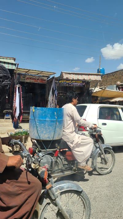 This media file captures a sunny street scene, likely a commercial area in Kuchlak, Pakistan. The image, oriented as if rotated 90 degrees counter-clockwise from its provided state, shows a clear blue sky with sparse white clouds and several parallel power lines stretching across the upper left.

The main subject is a man riding a motorcycle, seen from behind, wearing a light-colored traditional shalwar kameez and brown sandals. A large, weathered blue plastic barrel is strapped securely to the back of his motorcycle. His hands are on the handlebars, suggesting he is either riding or stopped in traffic. Another person's hands, wearing brown traditional clothing, are visible in the immediate foreground, holding the handlebars of what appears to be another motorcycle, partially obscuring the view of the first motorcycle's front wheel.

In the background, on the right side of the street, are several small shops or market stalls with striped awnings. A white car is parked behind the first man's motorcycle. The shops display various goods, possibly clothes or other general items, and their facades feature signage in Urdu script.

Notable details include the bright daylight indicated by strong shadows, the utilitarian nature of the motorcycles, and the presence of the blue barrel, which suggests the transport of liquids or other bulk goods. The architecture and clothing styles are consistent with the provided location context.

Visible text on the shop signs includes:
- "محمد عمر کیپ هاؤس" (Muhammad Omar Cap House)
- "محمد رضا کتب هاؤس" (Muhammad Raza Book House)
- Phone numbers: "0328.3881595" and "0308.6306969"
- "الکسی روایات" (Alaxi Riwayat)
On one of the motorcycle tires, the text "UNIQ", "MB-01", and "NYLON" can be seen.