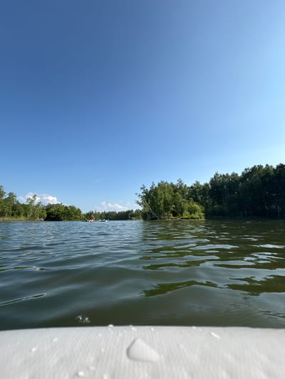 A bright, sunny day is depicted on a calm body of water, likely a lake or slow-moving river. The foreground shows the edge of a white object, possibly a boat or raft, with water droplets on its surface. The middle ground features the water with gentle ripples reflecting the green of the surrounding trees and the blue of the sky. In the distance, on the water, two people are visible on what appears to be an inflatable raft. They are positioned near the center of the frame. The background is dominated by lush green trees lining the banks of the water, with a clear blue sky overhead. A small island with trees is situated on the left side of the water. The scene suggests a recreational outing in a natural environment. The location is described as Razdolnoye, Russia.