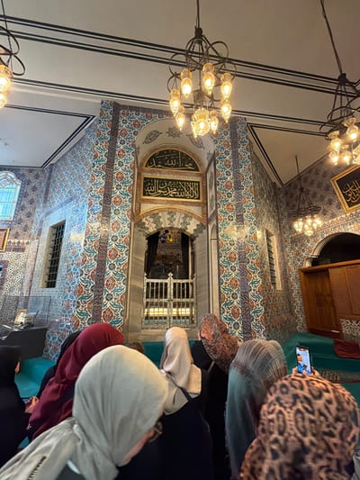 In the city of Eyüpsultan, Turkey, inside a sacred space adorned with intricate Iznik tiles and Islamic calligraphy, a group of individuals, primarily women wearing headscarves, are gathered. The architecture features arched doorways, ornate chandeliers, and latticed windows. A metal gate, possibly enclosing a tomb or sacred site, is visible in the background, illuminated by soft light. The individuals are standing, some are taking photographs with their mobile phones, indicating a moment of reverence or tourism. The overall atmosphere suggests a visit to a significant religious or historical landmark.