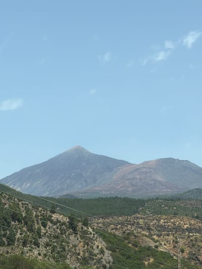 The image displays a dramatic mountain landscape under a clear blue sky with sparse white clouds. The mountainside is rugged and textured, with a mix of bare rock and dense green vegetation. In the foreground, several power lines stretch diagonally across the frame, suggesting the presence of human infrastructure within the natural setting. The mountains themselves are imposing, with steep slopes and a prominent peak dominating the right side of the image. The vegetation appears dry and hardy, typical of a Mediterranean climate. The lighting suggests it is daytime, likely mid-morning or afternoon, with the sun casting distinct shadows on the mountain faces. There is no visible text in the image. The scene evokes a sense of wilderness and the power of nature, with the power lines serving as a reminder of human presence and connectivity within this remote environment.