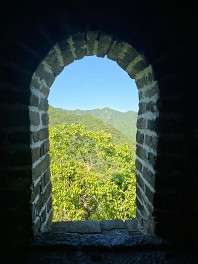 A view through an arched opening in a brick wall looks out onto a verdant mountainous landscape. The brickwork, composed of dark bricks with white mortar, forms a rough archway that frames the scene. The interior of the opening is dark, with the wall's shadow creating a strong contrast with the brightly lit exterior. Outside the arch, rolling green hills are covered in lush vegetation, with trees of varying shades of green and some lighter green foliage in the foreground. The sky above is a clear, bright blue. The opening appears to be a window or embrasure in a structure, possibly a historical fortification or watchtower, situated in a mountainous region of Huairou District, China. The foreground of the opening reveals a stone ledge made of rough-hewn stones.